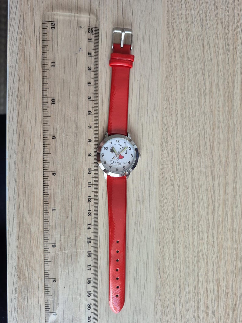 May include: A wristwatch with a bright red strap and a silver-tone case. The white watch face features a cartoon dog holding a red heart. A clear plastic ruler is placed alongside the watch, showing a total length of approximately 8.5 inches.