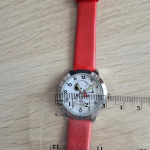 May include: A wristwatch with a bright red strap and a round silver-tone case. The white watch face features a graphic of Snoopy and black numerals. A clear plastic ruler is placed over the watch, showing a case diameter of approximately 1.25 inches.