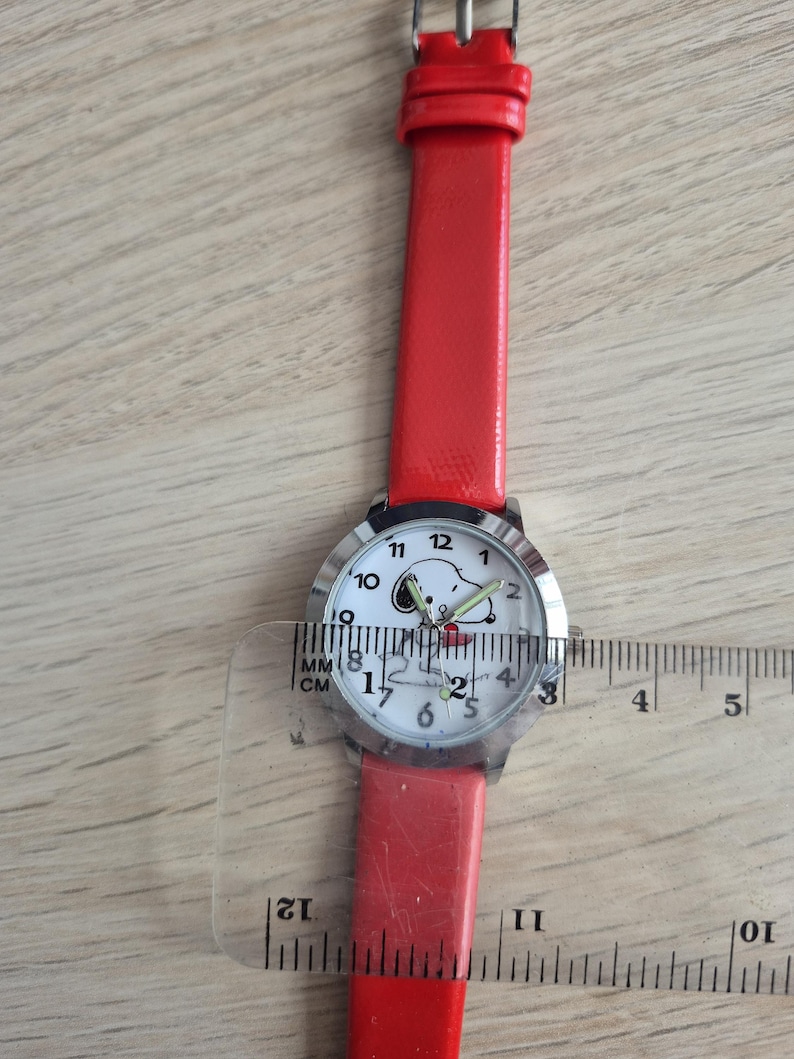 May include: A wristwatch with a bright red strap and a round silver-tone case. The white watch face features a graphic of Snoopy and black numerals. A clear plastic ruler is placed over the watch, showing a case diameter of approximately 1.25 inches.