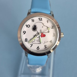 May include: A wristwatch with a light blue strap and a silver-tone metal case. The white watch face features black Arabic numerals, a graphic of a cartoon dog holding a red heart, and the signature Snoopy in black script near the number four.