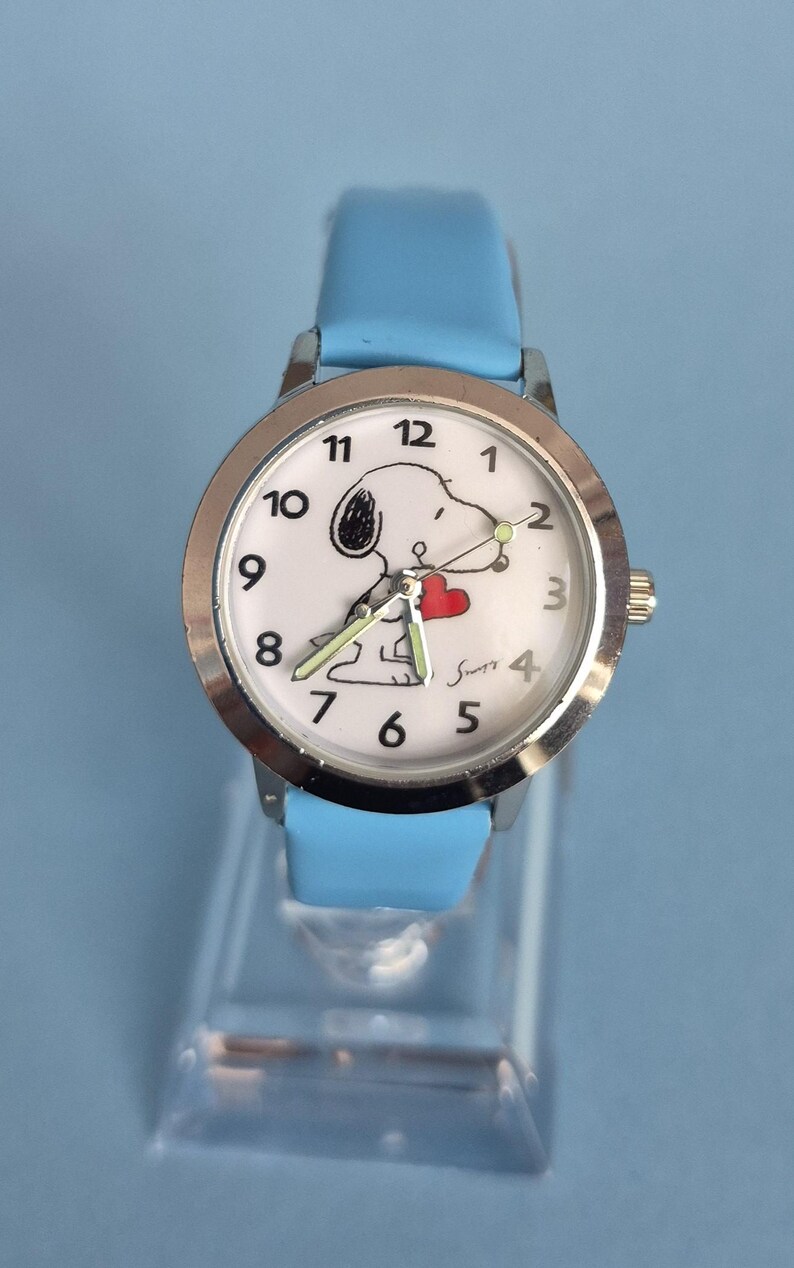 May include: A wristwatch with a light blue strap and a silver-tone metal case. The white watch face features black Arabic numerals, a graphic of a cartoon dog holding a red heart, and the signature Snoopy in black script near the number four.