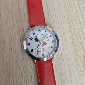 May include: A wristwatch with a glossy red strap and a silver-tone circular case. The white watch face features black Arabic numerals, a graphic of the character Snoopy holding a red heart, and the signature Snoopy in black script near the number 4.