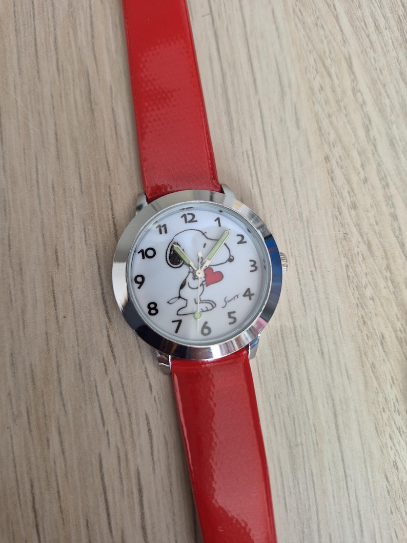 May include: A wristwatch with a glossy red strap and a silver-tone circular case. The white watch face features black Arabic numerals, a graphic of the character Snoopy holding a red heart, and the signature Snoopy in black script near the number 4.