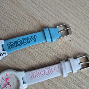 May include: Two Snoopy watches with light blue and white bands. The light blue band has the word "SNOOPY" in white. The white watch has a pink Snoopy face and the word "SNOOPY" in pink on the band.