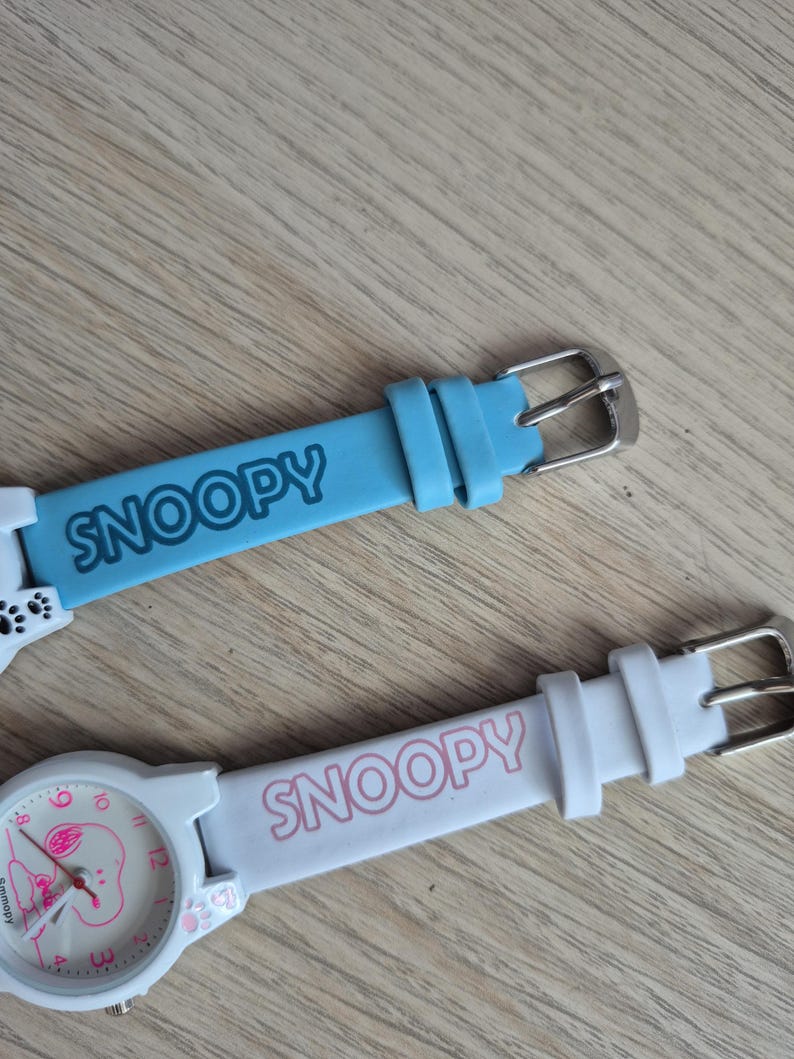 May include: Two Snoopy watches with light blue and white bands. The light blue band has the word "SNOOPY" in white. The white watch has a pink Snoopy face and the word "SNOOPY" in pink on the band.