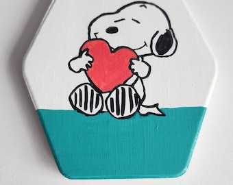 Puede incluir: Un posavasos hexagonal de madera blanco con fondo azul turquesa. Un perro de dibujos animados en blanco y negro está pintado en la parte superior sosteniendo un corazón rojo.