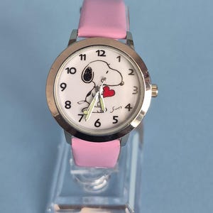May include: A wristwatch with a silver-tone metal case and a light pink leather strap. The white watch face features black numerals, a graphic of the character Snoopy holding a red heart, and three hands. The name Snoopy is printed in black.
