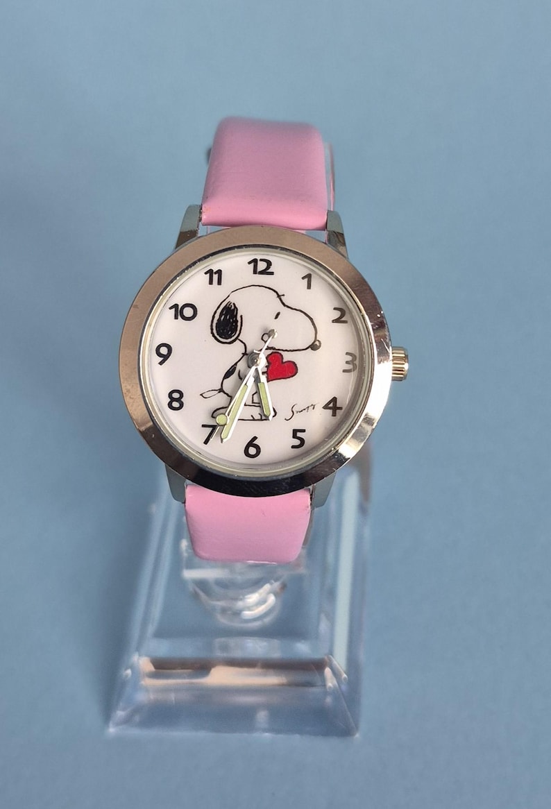 May include: A wristwatch with a silver-tone metal case and a light pink leather strap. The white watch face features black numerals, a graphic of the character Snoopy holding a red heart, and three hands. The name Snoopy is printed in black.