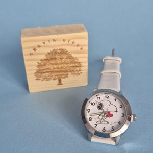 May include: A wristwatch with a white strap and a silver-tone bezel featuring a white dial with black numerals and a graphic of Snoopy holding a red heart. Beside the watch is a square wooden block engraved with a tree and the text CHEVIN GIFTS.