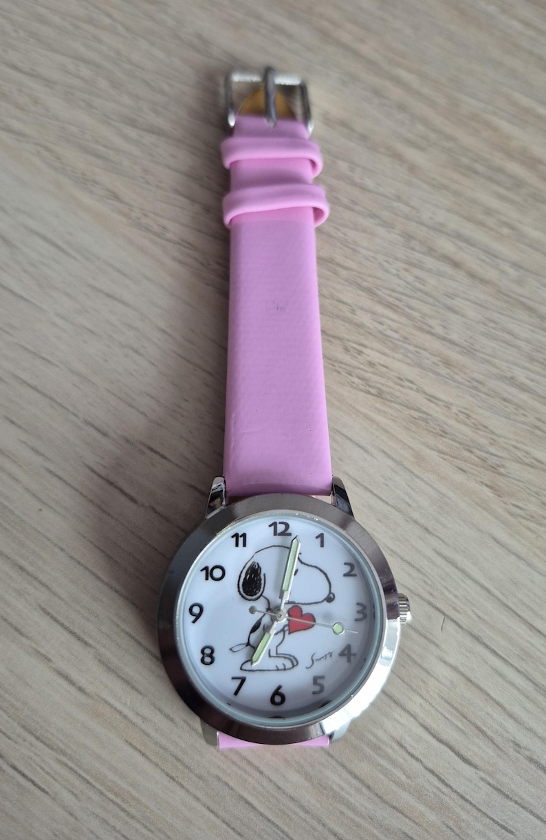May include: A wristwatch with a pink strap and a silver-tone round case. The white watch face features black numerals, a graphic of a cartoon dog holding a red heart, and the signature Snoopy. The watch has silver-tone hands and a buckle closure.