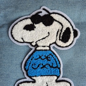 May include: Embroidered patch of Snoopy wearing sunglasses and a blue sweater. The patch has a white background with black details and a light blue border. The design is in a cartoon style.