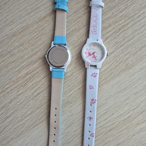 May include: Two wristwatches. One has a light blue leather strap and a silver-colored watch face. The other watch has a white strap with pink paw prints and the word "SNOOPY". The watch face features Snoopy.
