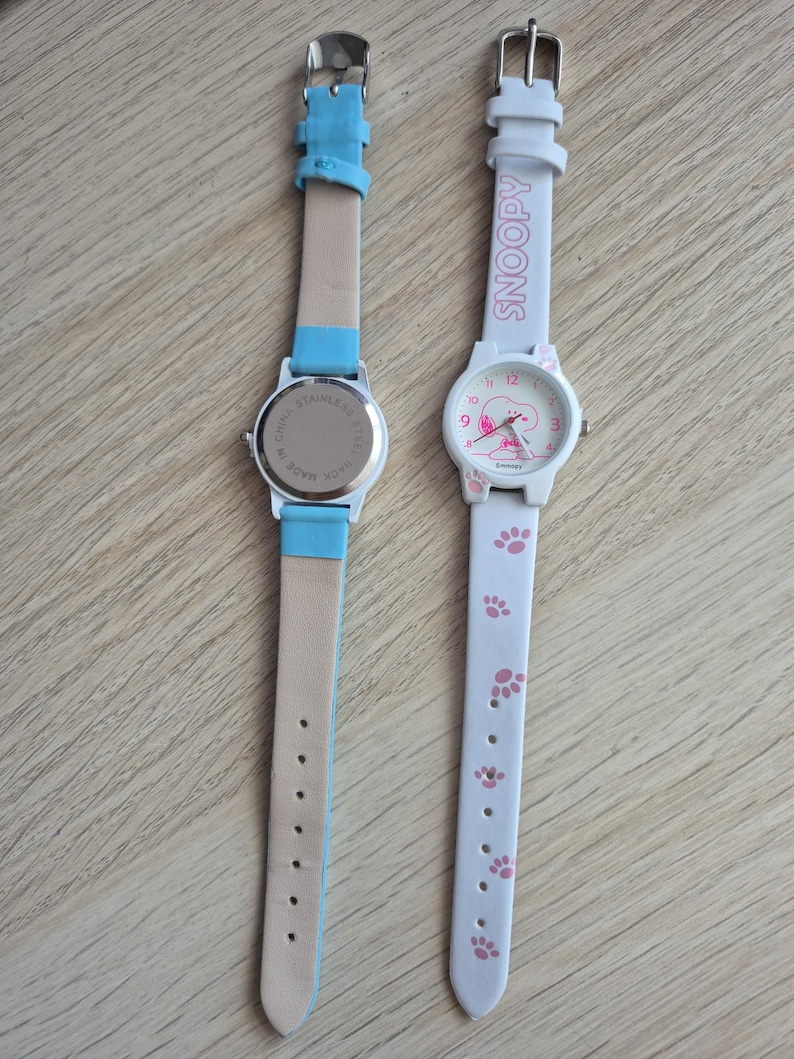 May include: Two wristwatches. One has a light blue leather strap and a silver-colored watch face. The other watch has a white strap with pink paw prints and the word "SNOOPY". The watch face features Snoopy.