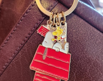 May include: A Snoopy keychain with a red doghouse charm and a gold-colored keyring. The charm features Snoopy and Woodstock in a red doghouse. The keychain is attached to a purple leather purse.