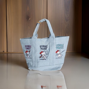 Op de afbeelding: Lichtblauwe denim tote bag met Snoopy honkbal illustraties. De tas toont Snoopy in honkbal poses met de tekst "Strike One!", "Strike Two!" en "Strike Three!". Het heeft een handvat met een houten accent. Geschikt voor kleine items.