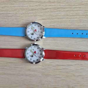 Op de afbeelding: Twee polshorloges met Snoopy-wijzerplaten. Eén horloge heeft een lichtblauwe band, de andere een rode band. Beide horloges hebben zilverkleurige kasten en gespen. De wijzerplaten tonen Snoopy met een hart.