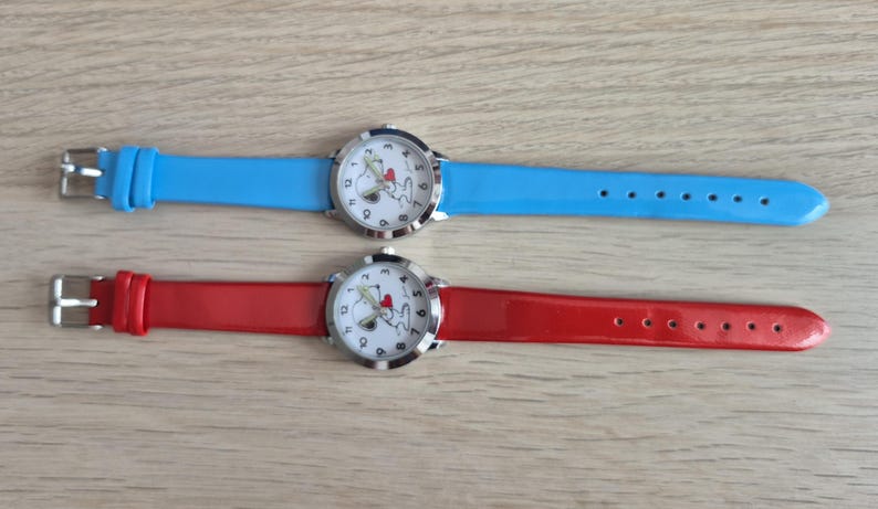 May include: Two analog wristwatches with round silver-tone cases and white dials featuring a cartoon dog graphic with a red heart. One watch has a bright blue strap, and the other has a vibrant red strap, both with silver-tone metal buckles.