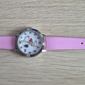 May include: A wristwatch with a light pink strap and a silver-tone metal case. The white watch face features black Arabic numerals and a graphic illustration of a cartoon dog holding a red heart. The watch includes a silver buckle and loops.