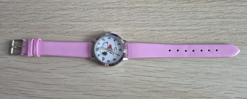 May include: A wristwatch with a light pink strap and a silver-tone metal case. The white watch face features black Arabic numerals and a graphic illustration of a cartoon dog holding a red heart. The watch includes a silver buckle and loops.
