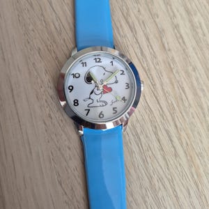 May include: A wristwatch with a bright blue strap and a silver-tone metal case. The white watch face features black Arabic numerals and a graphic illustration of the character Snoopy holding a small red heart, with silver-tone hour and minute hands.