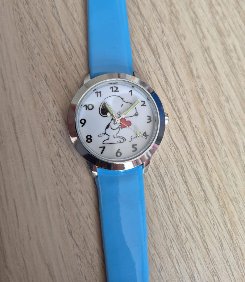 May include: A wristwatch with a bright blue strap and a silver-tone metal case. The white watch face features black Arabic numerals and a graphic illustration of the character Snoopy holding a small red heart, with silver-tone hour and minute hands.