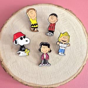 以下が含まれることがあります： 漫画のキャラクターが描かれた5つのエナメルピンのセット。白い犬と赤い帽子のピン、黄色いシャツを着たキャラクター、赤いジャケット、ピンクのジャケット、そして「Gucci」という文字が入った白いシャツを着たキャラクターのピンが含まれています。