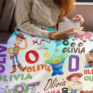 May include: A soft blanket featuring a colorful Toy Story character print with Woody, Buzz Lightyear, Jessie, Mr. Potato Head, and an alien. The design includes the name Olivia in various fonts and colors, alongside stars, flowers, and letters.
