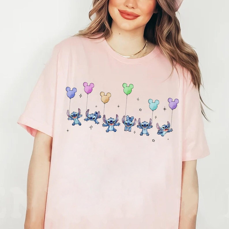 May include: A light pink t-shirt featuring a cartoon design of Stitch characters holding colorful balloons. The balloons are in shades of blue, pink, yellow, and green. The design is centered on the front of the shirt.
