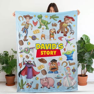 May include: A light blue fleece blanket featuring a colorful print of various animated characters and objects from a popular film series, including a cowboy, space ranger, dinosaur, and potato figure, with the text DAVID'S STORY in the center.