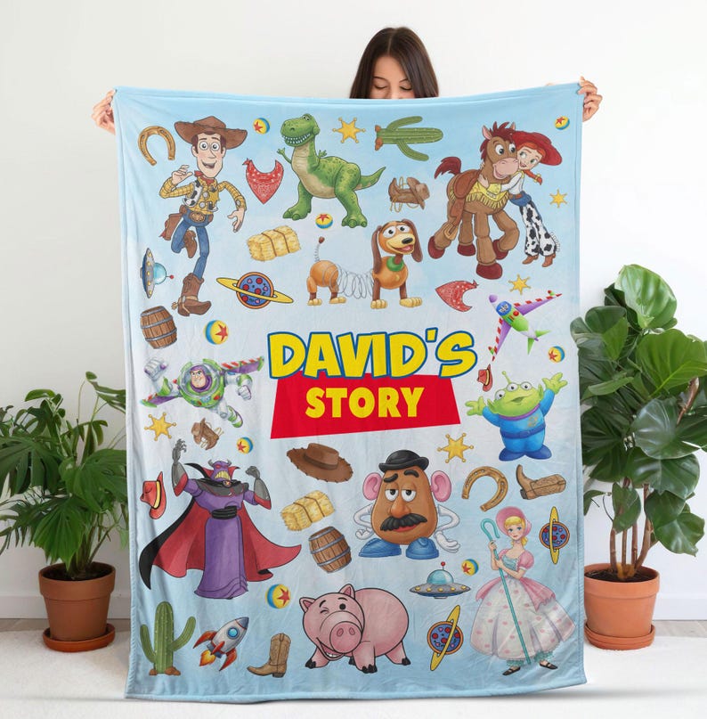 May include: A light blue fleece blanket featuring a colorful print of various animated characters and objects from a popular film series, including a cowboy, space ranger, dinosaur, and potato figure, with the text DAVID'S STORY in the center.