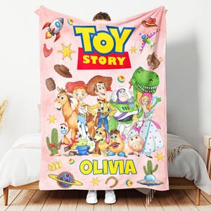 May include: A light pink fleece blanket featuring the Toy Story logo at the top and the name OLIVIA printed in yellow at the bottom. The blanket displays various characters including Woody, Buzz Lightyear, Jessie, Rex, and others with icons.