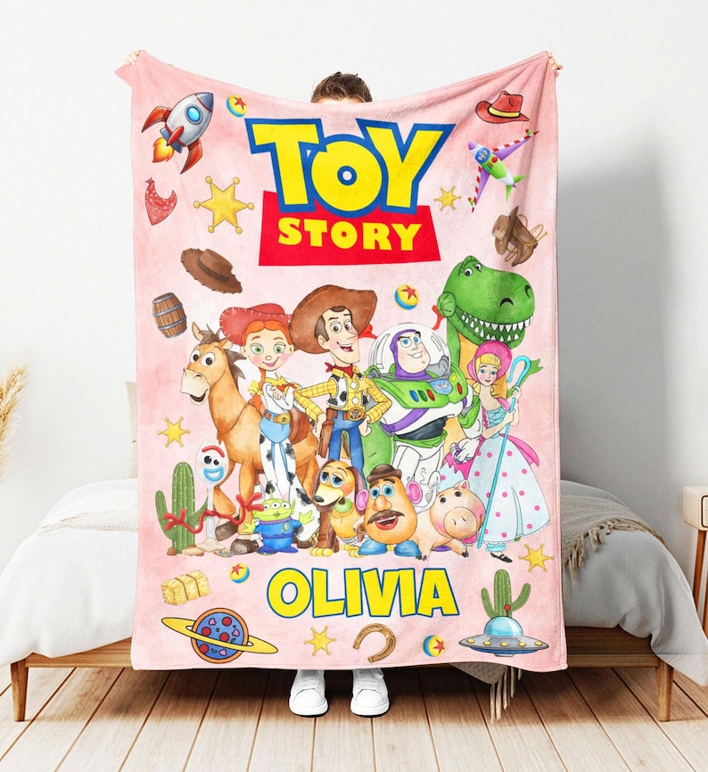 May include: A light pink fleece blanket featuring the Toy Story logo at the top and the name OLIVIA printed in yellow at the bottom. The blanket displays various characters including Woody, Buzz Lightyear, Jessie, Rex, and others with icons.