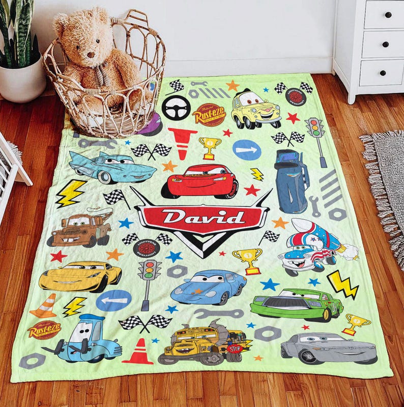 May include: A light green fleece blanket featuring a colorful pattern of various cartoon cars, traffic lights, checkered flags, and trophies. The center displays a red emblem with the name David in white text, surrounded by scattered stars.