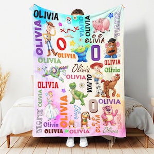May include: A soft fleece blanket featuring a colorful watercolor background with various cartoon characters including a cowboy, space ranger, cowgirl, dinosaur, and pig. The name OLIVIA is printed repeatedly in various fonts, colors, and sizes.
