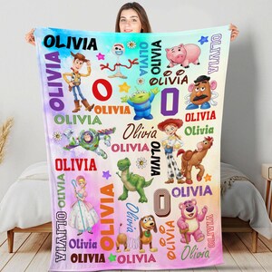 May include: A rectangular fleece blanket featuring a colorful watercolor background with various Toy Story character illustrations including Woody, Buzz Lightyear, Jessie, Rex, and Hamm. The name Olivia is printed repeatedly in various fonts and colors.
