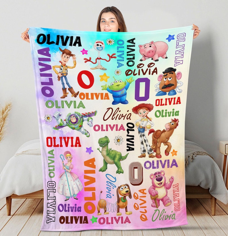 May include: A rectangular fleece blanket featuring a colorful watercolor background with various Toy Story character illustrations including Woody, Buzz Lightyear, Jessie, Rex, and Hamm. The name Olivia is printed repeatedly in various fonts and colors.