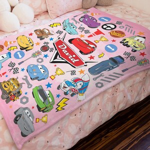 May include: A soft pink fleece blanket featuring a repeating pattern of various cartoon race cars, checkered flags, traffic lights, wrenches, and gold trophies. A large red emblem in the center displays the name David in white cursive lettering.