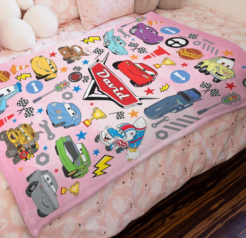 May include: A soft pink fleece blanket featuring a repeating pattern of various cartoon race cars, checkered flags, traffic lights, wrenches, and gold trophies. A large red emblem in the center displays the name David in white cursive lettering.