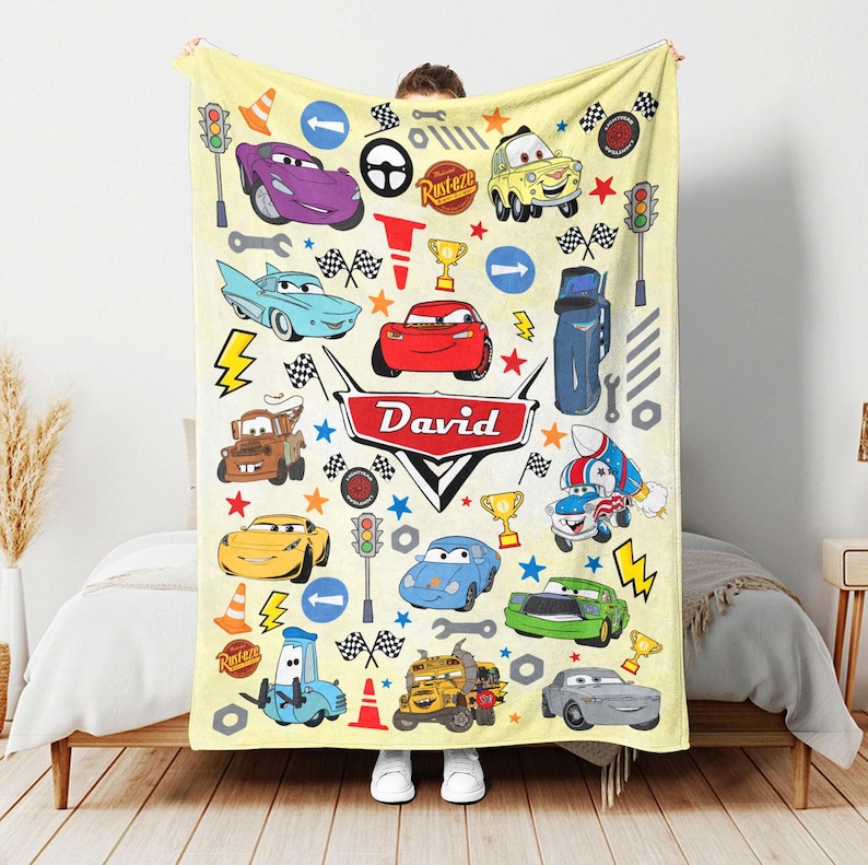 May include: A light yellow fleece blanket featuring a repeating pattern of various characters and icons from the movie Cars, including Lightning McQueen, Mater, and others, with the name David printed in the center on a red emblem.