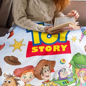 May include: A white fleece blanket featuring the Toy Story logo in blue and yellow text on a red background. The print includes illustrations of Woody, Jessie, Buzz Lightyear, Rex, Bo Peep, Bullseye, a cowboy hat, a rocket, and yellow stars.