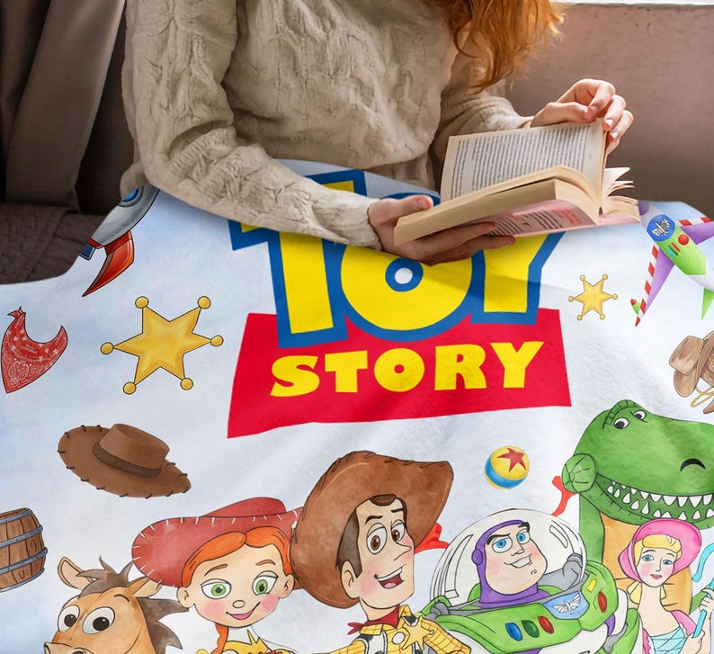 May include: A white fleece blanket featuring the Toy Story logo in blue and yellow text on a red background. The print includes illustrations of Woody, Jessie, Buzz Lightyear, Rex, Bo Peep, Bullseye, a cowboy hat, a rocket, and yellow stars.