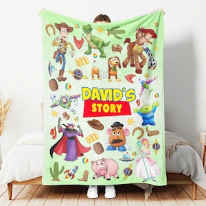 May include: A light green fleece blanket featuring various Toy Story character illustrations, including Woody, Buzz Lightyear, Rex, Slinky Dog, and Mr. Potato Head, with the text "DAVID'S STORY" printed in large yellow and red letters in the center.