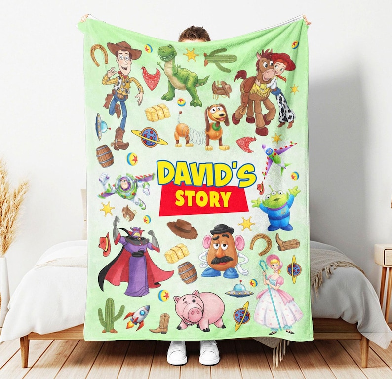 May include: A light green fleece blanket featuring various Toy Story character illustrations, including Woody, Buzz Lightyear, Rex, Slinky Dog, and Mr. Potato Head, with the text "DAVID'S STORY" printed in large yellow and red letters in the center.