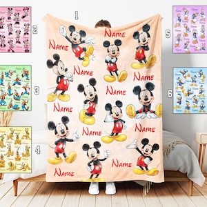 Puede incluir: Una manta de forro polar suave con un patrón repetitivo de Mickey Mouse en varias posturas sobre un fondo color melocotón claro, intercalado con el texto rojo "NAME" varias veces. Seis variaciones más pequeñas muestran otros personajes.