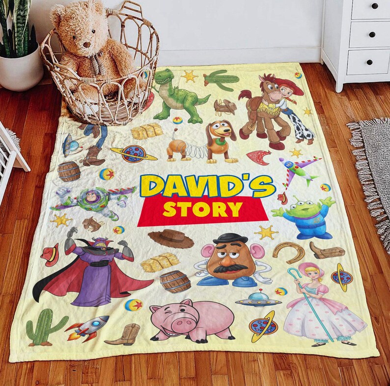 May include: A light yellow fleece blanket featuring various Toy Story characters including Buzz Lightyear, Woody, Jessie, Bullseye, Rex, Slinky Dog, Mr. Potato Head, Bo Peep, and Zurg, with the text DAVID'S STORY printed in large blue and red letters.