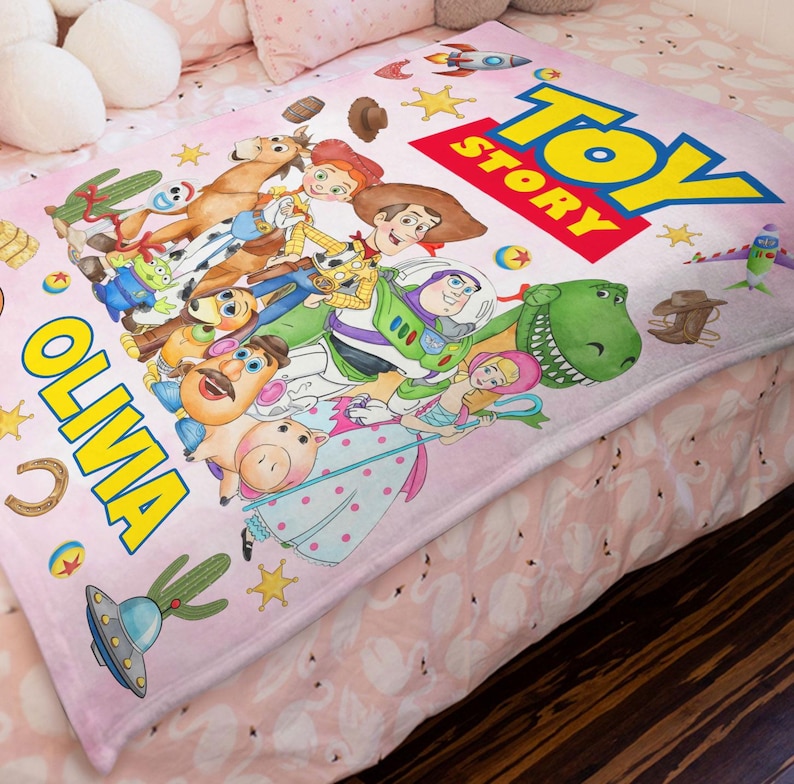 May include: A soft pink fleece blanket featuring the Toy Story logo and characters including Woody, Buzz Lightyear, Jessie, Rex, and Mr. Potato Head. The blanket is personalized with the name OLIVA in large yellow letters and decorated with stars, cacti, and space-themed icons.