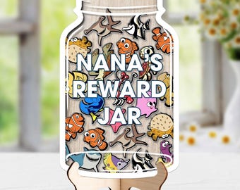 Könnte beinhalten: Ein holzfarbenes, glasförmiges Schild mit einer Sammlung bunter Cartoon-Meeresbewohner-Illustrationen und dem weißen Text NANA'S REWARD JAR. Das Schild wird von einem hellen Holzständer gehalten und zeigt verschiedene Fische und Meerestiere.