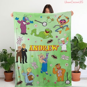 Puede incluir: Una manta de forro polar verde claro con varios personajes de los Muppets, incluyendo a la rana Gustavo, la cerdita Peggy, Gonzo y el oso Fozzie. La manta muestra el nombre ANDREW en letras amarillas grandes, entre flores blancas y estrellas.