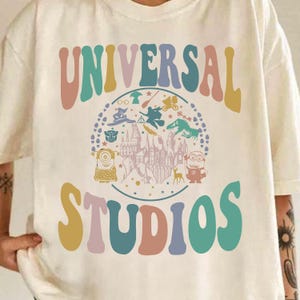 May include: Cream-colored t-shirt featuring the words "UNIVERSAL STUDIOS" in a retro font. The shirt has a circular graphic with various Universal Studios characters and icons, including Minions, a dragon, and a castle.