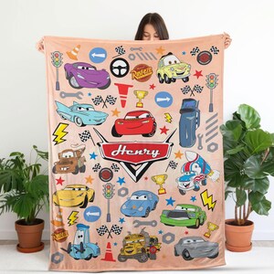May include: A light peach-colored fleece blanket featuring a repeating pattern of various cartoon cars, traffic lights, checkered flags, and tools from the movie Cars. The center displays a large red emblem with the name Henry written in white.